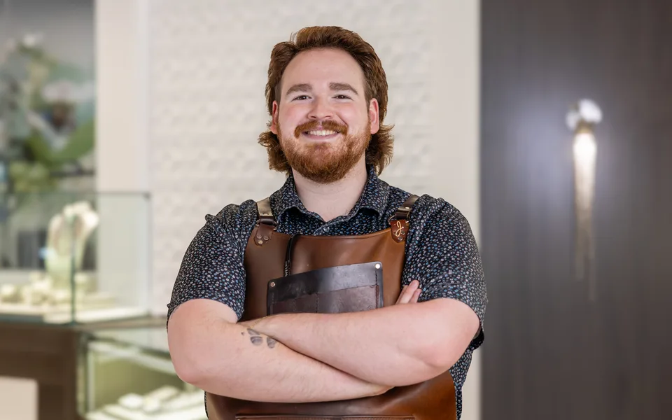 One of our goldsmiths in a leather bench apron, arms folded, smiling in the showroom